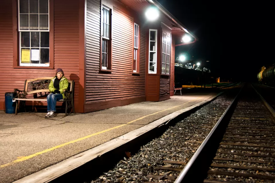 Womanin yellow jacket sitting on bench at outdoor train station with red wood depot building at night.
